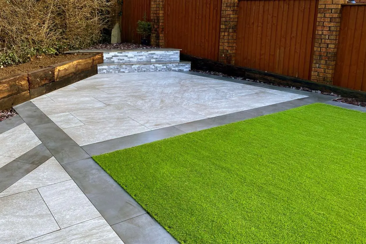 A modern backyard with a neatly landscaped design features large, light gray tiled patio sections bordered by darker tiles and a lush, bright green waterless lawn from Coachella Valley Turf. Wooden fencing and a set of steps leading upwards are seen in the background, reminiscent of Palm Springs elegance.