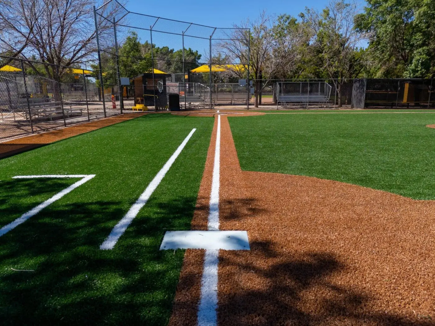Two gardening gloves lie on a vibrant green lawn reminiscent of a well-maintained sports field astroturf. One glove is slightly closer to the foreground, with both gloves showing a mix of purple, yellow, and white colors. The scene is brightly lit, highlighting the gloves against the lush grass background.
